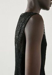 Robe en dentelle noire avec un design sans manches, texture à mailles ouvertes et encolure ronde, présentée sous un angle latéral.