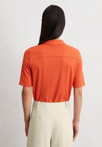 Oranje poloshirt met een detail op de achterkraag, gecombineerd met lichtbeige hoge taille broeken met twee achterzakken.