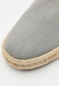 Espadrille en toile grise avec un bout arrondi, semelle en corde de jute et surface en tissu texturé. Présente des coutures simples le long du bord.