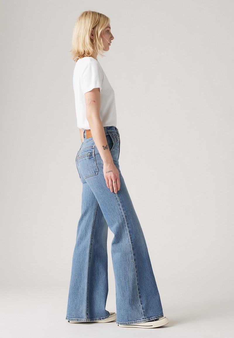 Jean évasé bleu clair en denim, avec une taille haute, des coutures contrastées et un détail de poche zippée sur le côté.