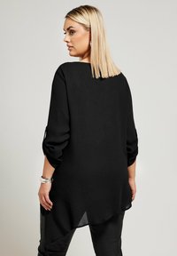 Maglietta nera a maniche lunghe con una vestibilità ampia, caratterizzata da un orlo arrotondato e maniche leggermente arrotolate. Nessun motivo o accessorio visibile.