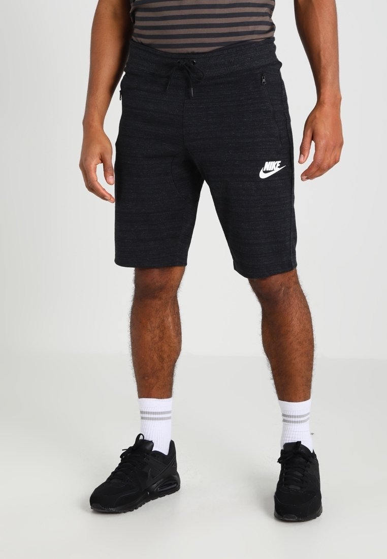 Homme portant un short noir Nike avec poches zippées, des baskets noires, des chaussettes blanches avec des rayures grises et une chemise rayée coupée à la taille.