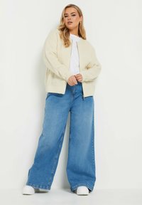 Krämfärgad stickad cardigan med en avslappnad passform, ribbad yta och långa ärmar, kombinerad med ljusblå vida jeans och vita sneakers.