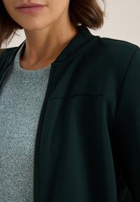 Mörkgrön bomberjacka med en slät textur, med dragkedja framtill och en ljusgrön fläckig tröja under, synlig krage.