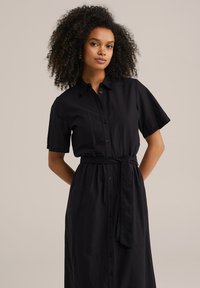 Abito camicia nero con maniche corte, realizzato in tessuto leggero, caratterizzato da una chiusura frontale con bottoni e una vita legata. Texture liscia, design casual.
