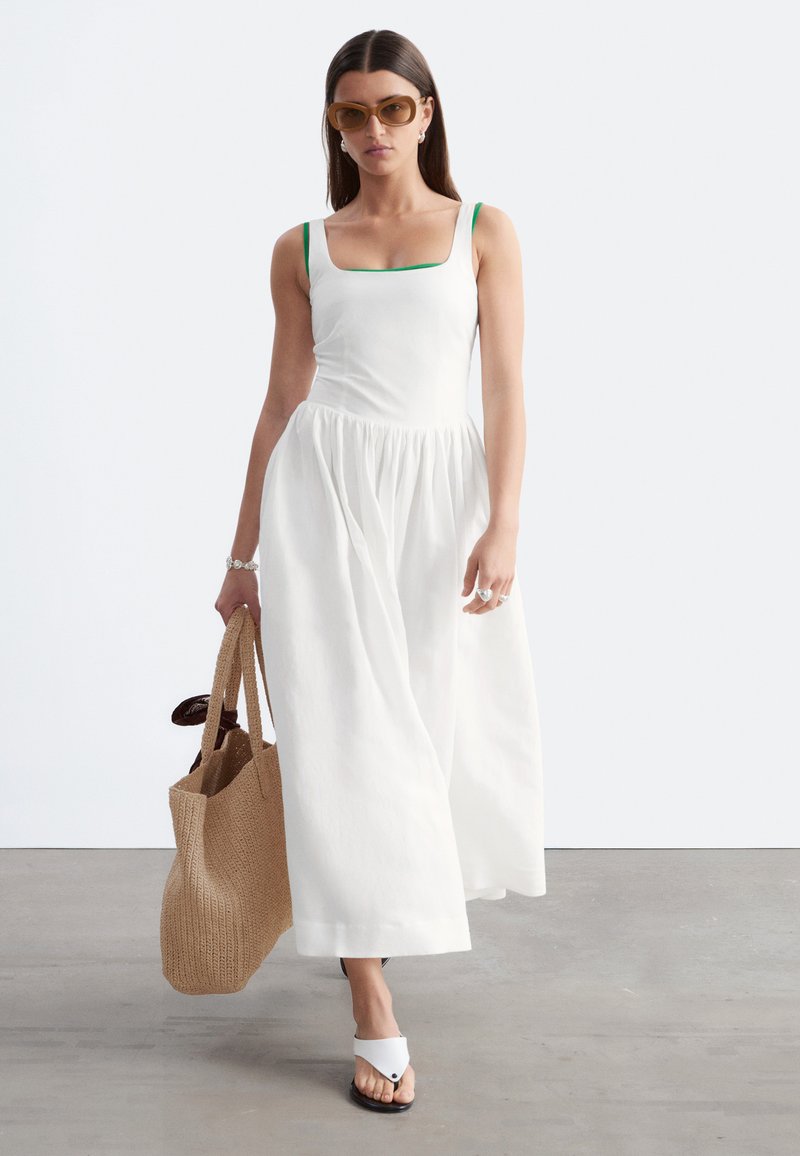 Femme aux cheveux bruns et lisses portant une robe blanche sans manches et des lunettes de soleil marron, portant un grand sac cabas tissé, marchant sur un sol gris.