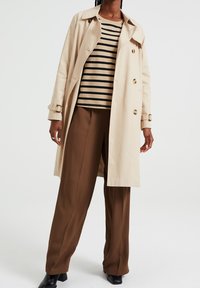 Osoba v béžovém trenčkotě přes světlehnědém a černém pruhovaném tričku, s hnědými širokými kalhotami a černými vysokými botami na podpatku na bílém pozadí.