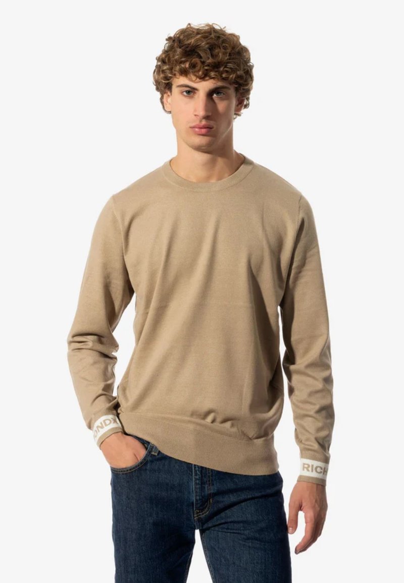 Suéter de algodón beige con mangas largas, cuello redondo y puños de canalé con acentos de logo blanco. Combinado con jeans azules.