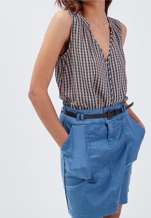 Woman wearing a sleeveless patterned blouse tucked into a belted blue skirt with hands in pockets against plain background.