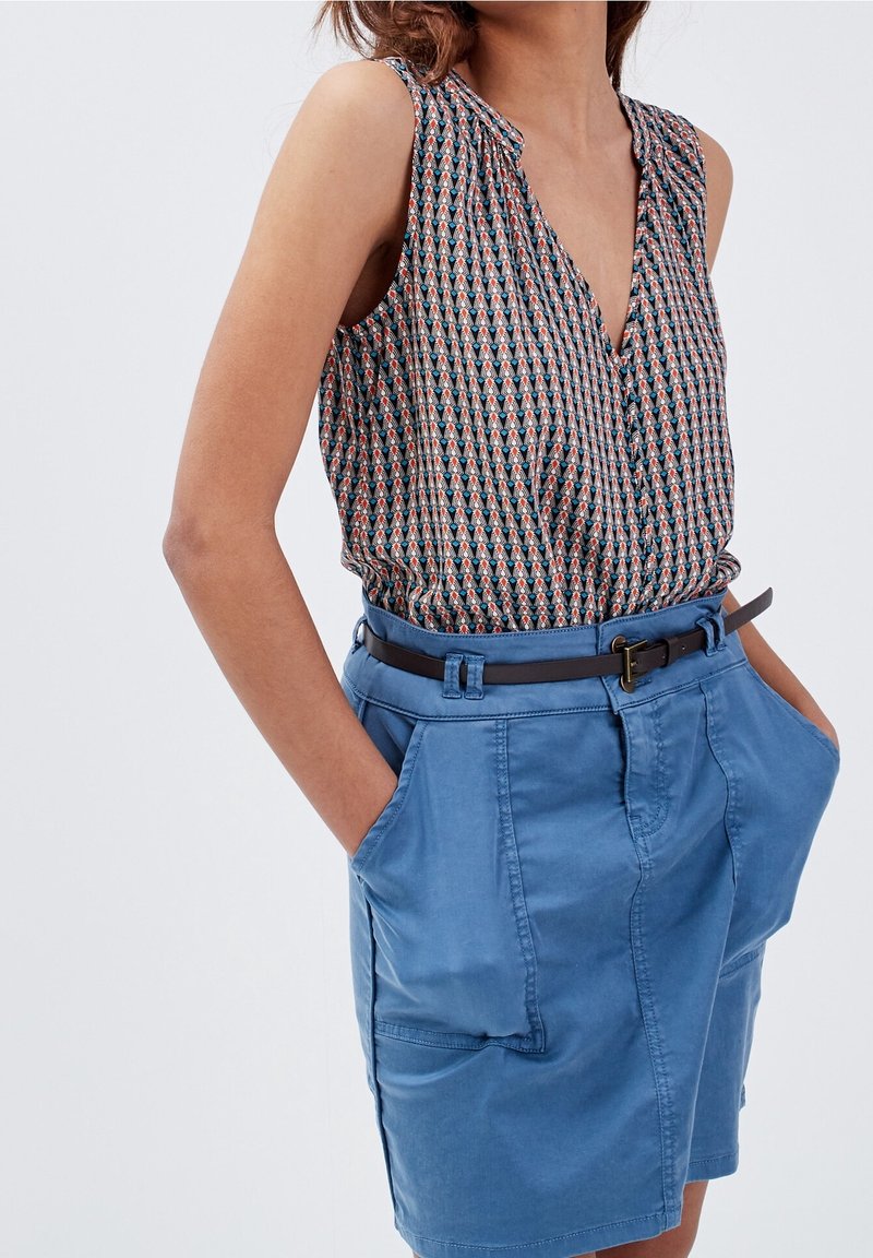 Femme portant un chemisier sans manches à motifs, rentré dans une jupe bleue ceinturée, les mains dans les poches, sur un fond uni.