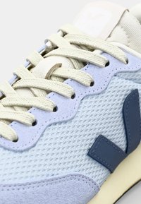 Sapatilha azul clara com um cabedal de tecido texturizado, um logótipo azul marinho liso, cilindros em creme e um puxador de calcanhar beige arredondado.