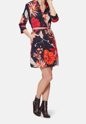 Femme portant une robe bleu marine avec de grands motifs floraux rouges et orange, des manches trois-quarts, une ceinture à la taille et des bottines marron foncé.