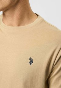 Közelkép egy bézs pólóról, amelyen sötétkék hímzett polo játékos logo látható a mellkason, és egy férfi nyaka és állvonala is látszik.