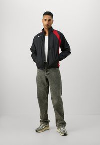 Veste coupe-vent noire et rouge avec fermeture éclair sur le devant, poignets élastiques et un petit logo. Portée avec un jean gris et des baskets épaisses.