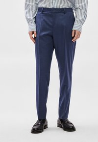 Παντελόνι φόρεμα σε ναυτικό μπλε με ομαλή υφή και εφαρμοστή γραμμή. Διαθέτει πιέτες μπροστά και θηλιές για ζώνη· συνδυασμένο με μαύρα μοκασίνια.