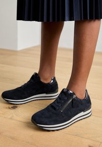 Ben som bär en plisserad marinblå kjol och svarta mockaskor med sidodetalj, står på trägolv.