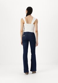 White smocked top with wide straps paired with dark blue flared jeans featuring back pockets and a logo tag. Smooth fabric texture.