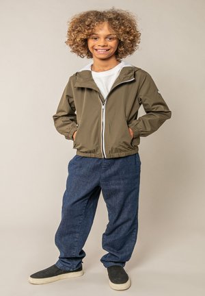Enfant souriant aux cheveux bouclés portant une veste zippée vert olive, un t-shirt blanc, un jean ample bleu et des chaussures noires sans lacets, debout devant un mur beige.