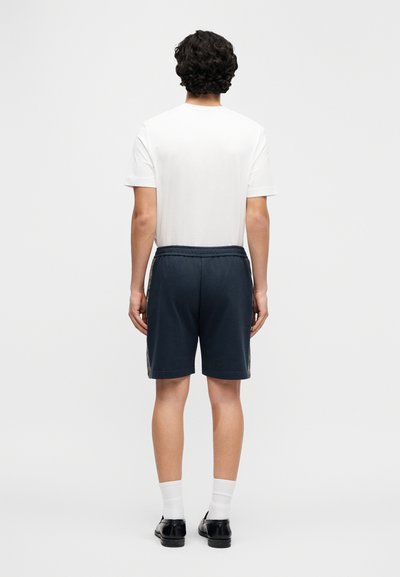Λευκό T-shirt σε συνδυασμό με σκούρο μπλε σορτς που διαθέτει υφασμάτινη πλευρική λεπτομέρεια, ελαστική μέση και μήκος πάνω από το γόνατο.