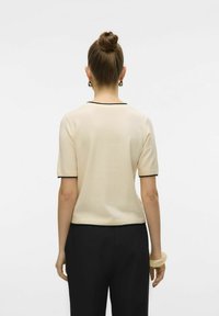 En ung kvinna med en knut i håret, iklädd en krämfärgad kortärmad topp med svart kant, vänder ryggen mot kameran och framhäver sin enkla, eleganta stil.