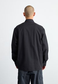 Zwarte longsleeve shirt met een kraag, met een gladde textuur en een ontspannen pasvorm, gezien vanaf de achterkant met knoopmanchetten.