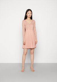 Vestido rosa com corpete de renda e mangas longas, saia fluida, decote em V e textura suave. Modelo em pé sobre um fundo neutro.