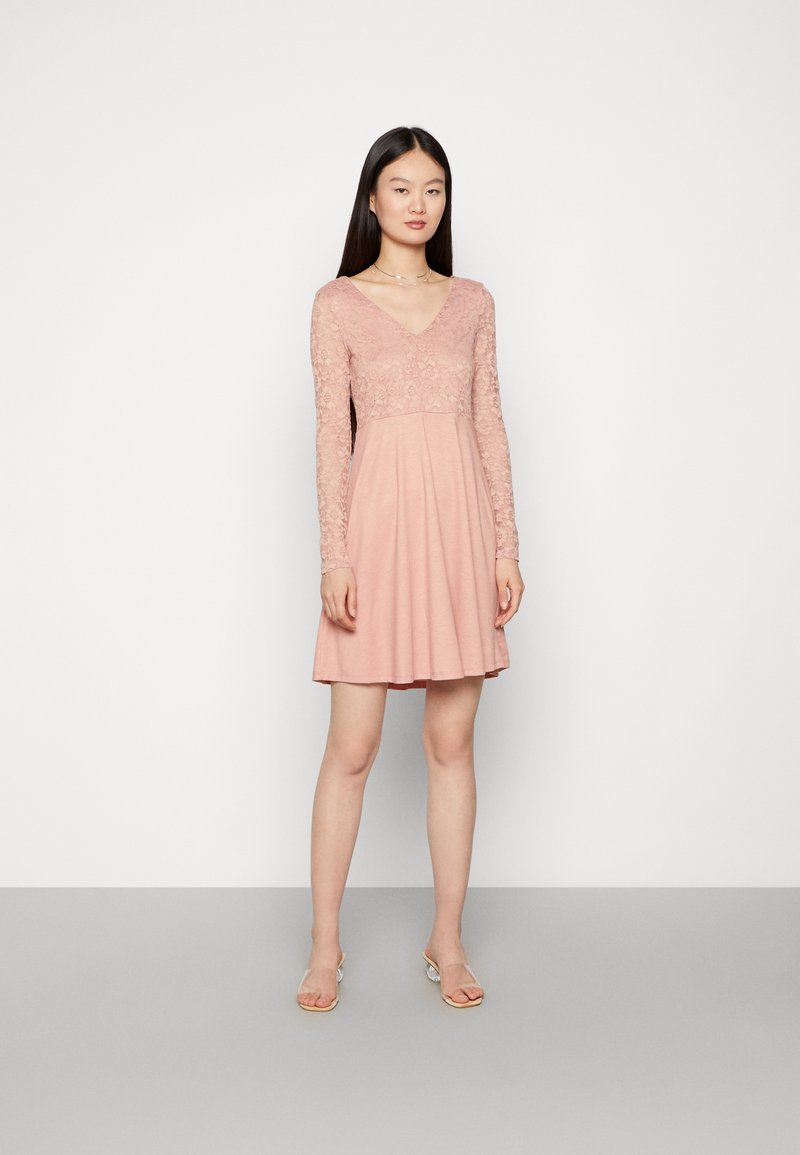 Vestido rosa com corpete de renda e mangas longas, saia fluida, decote em V e textura suave. Modelo em pé sobre um fundo neutro.