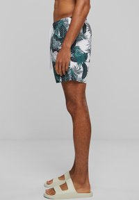 Shorts de bain avec une base blanche et un motif de feuilles vertes tropicales, dotés d'une taille élastique avec cordon de serrage. Portés avec des sandales de couleur claire.