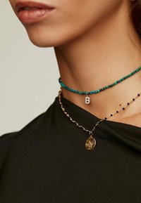 Collares de oro y turquesa superpuestos en un cuello, con un diseño de cuentas y un colgante con un emblema decorativo. Parte superior de tela negra texturizada.