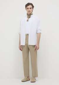 Uomo che indossa una camicia bianca con bottoni sopra una maglietta bianca, pantaloni beige, scarpe slip-on beige e una sciarpa a motivi marrone chiaro attorno al collo.