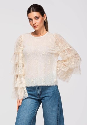 Mujer con blusa de encaje color crema con mangas abullonadas en capas y jeans azules, posando con la mano en la cadera frente a un fondo liso.