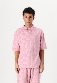 Short-sleeve, pink polo shirt with a textured pattern of squares and stylized motifs, made of soft fabric, featuring a buttoned collar.