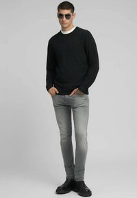 Zwarte trui met een ronde halslijn, grijze slim-fit jeans en zwarte enkellaarzen. Model draagt een donkere zonnebril en staat tegen een neutrale achtergrond.