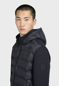 Gilet imbottito nero con cappuccio, design trapuntato e chiusura con zip. Indossato sopra una maglia nera a maniche lunghe, con un patch logo sulla manica.