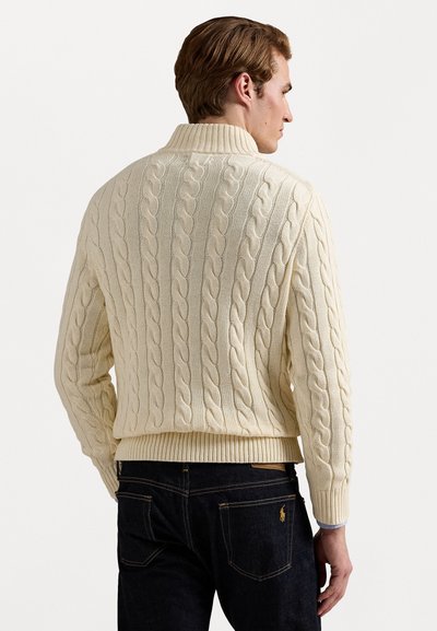 Kremasti pulover s pletenicom i visokim ovratnikom, s rebrastim rukavima i rubovima, uparen s tamnim trapericama koje imaju suptilno zlatno brendiranje na stražnjem džepu.