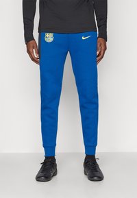Calças de jogging azuis, fabricadas em tecido suave, com um logotipo elevado do Barcelona em amarelo na coxa esquerda e um pequeno swoosh da Nike em dourado.