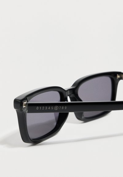 Κοντινό πλάνο σε μαύρα ορθογώνια γυαλιά ηλίου με σκούρους φακούς και αριθμητικό λογότυπο χαραγμένο στον κίονα που δείχνει ψηφία από 0 έως 9 με το 6 κυκλωμένο.