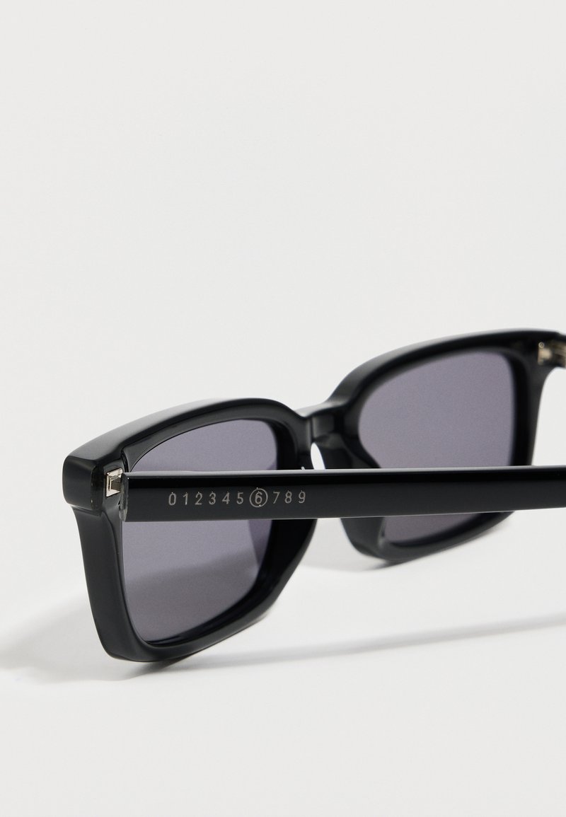 Primo piano di occhiali da sole rettangolari neri con lenti scure e un logo numerico inciso sulla stanghetta che mostra le cifre da 0 a 9 con il 6 cerchiato.