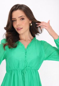 Rochie verde cu decolteu în formă de inimă, mâneci lungi, talie cu cordon și un material texturat, ușor. Prezintă un guler plisat.