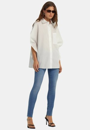 Donna che indossa una camicia bianca oversize con bottoni, jeans skinny blu, sandali neri con tacco alto e occhiali da sole rettangolari scuri, in piedi su sfondo bianco.