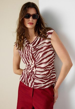 Frau mit langen braunen Haaren, die dunkle, rechteckige Sonnenbrillen trägt, ein ärmelloses, zebra-gestreiftes Oberteil und rote Hosen, stehend vor einer hellen Wand.