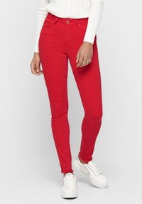 Jeans ajustados rojos con una textura suave, diseño de cinco bolsillos y un dobladillo deshilachado, combinados con zapatillas blancas y un suéter blanco de punto.