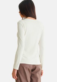 Langsleeve, ribbeltop in off-white stof met een ronde halslijn, getailleerde silhouet en een gladde textuur, in combinatie met bruine broek.