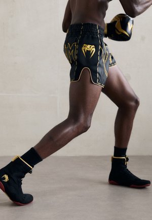 Atleta con pantalones cortos de boxeo negros y amarillos y guantes, llevando zapatillas de boxeo de caña alta negras y rojas, posicionado en una postura de pelea.