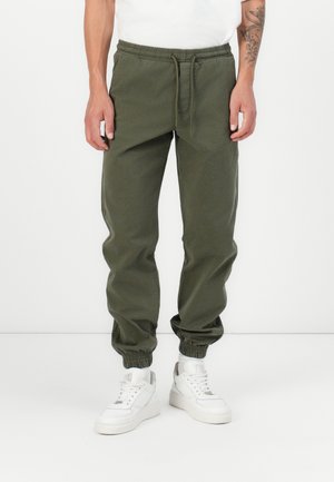 Joggers vert olive en tissu doux, dotés d'une taille élastique avec cordon de serrage, de jambes fuselées et de poignets élastiques. Associés à des baskets blanches.