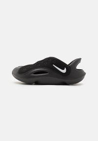 Mustat urheilu-slip-on -kengät, joissa on teksturoitu pinta, läpivientireiät hengittävyyden lisäämiseksi ja valkoinen swoosh-logo sivussa. Kaareva kantapään muotoilu.