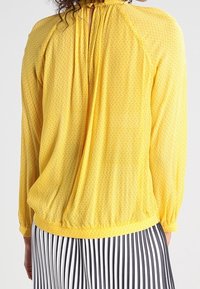 Blouse jaune à manches longues avec un motif texturé, dos froncé, poignets élastiques et coupe loose ; portée avec un pantalon à rayures noir et blanc.