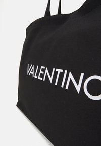 Svart kanvastotepåse med stor vit text "VALENTINO". Den har kraftiga axelremmar och en enkel, funktionell design.