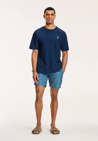 Tamnoplava majica kratkih rukava s tirkiznim logotipom, u kombinaciji s plavim kupaćim gaćama s uzorkom i laganim sandalama. Ležerna odjeća za plažu.