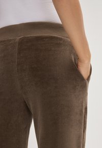 Bruine velours joggers met een gladde textuur, voorzien van een rekbare tailleband en zijzakken. Dichtbijzicht van de achterkant en het detail van de zak.
